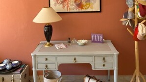 Ein Zimmer mit weißem Schreibtisch, einer Lampe, einem gerahmten abstrakten Gemälde, einem Haken für Hüte und alten Koffer.