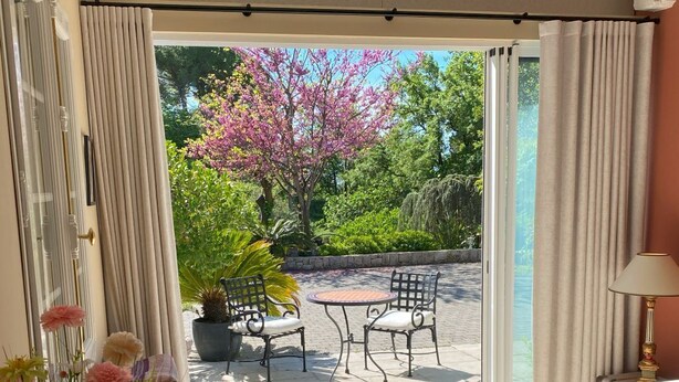 Ein Balkon mit Bett, Tisch, Stühlen, einem Blumentopf und Blick auf Bäume und einen Garten.
