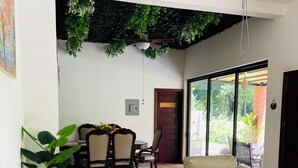 A modern living room with a beige sofa, patterned cushions, a ceiling fan, and a view of the outdoors through large windows.