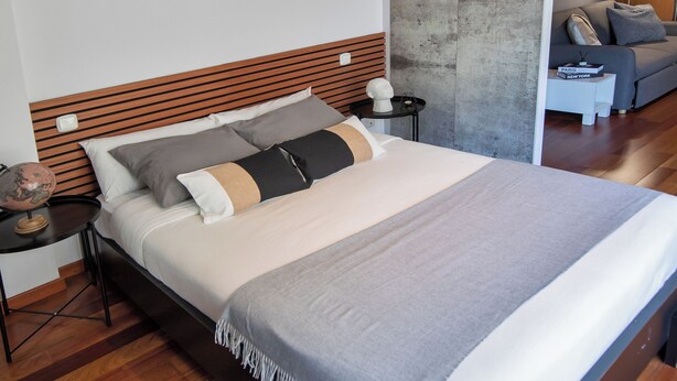 Ein modernes Schlafzimmer mit einem Holzkopfteil, einem großen Bett mit vielen Kissen, einem Nachttisch mit Lampe und einer Wand mit Betonoptik.