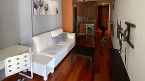 Ein modernes Wohnzimmer mit einem weißen Sofa, einem kleinen weißen Tisch und einem schwarzen Couchtisch. An der Wand hängt ein Gemälde und im Hintergrund steht eine Waschmaschine.
