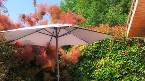 Une terrasse extérieure avec une table et deux chaises, un grand parasol, et un décor composé de feuillage et d’un bâtiment.