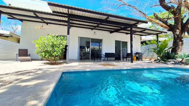 A modern outdoor pool area with a covered patio, seating, and a small garden.