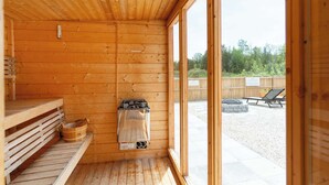 A wooden sauna room with a sauna heater, a bench, and a window leading to an outdoor area with a barbecue grill and lounge chairs. - Grand Luxe Geodome Glamping w/ 2 Queen beds East (Miller Lake)
