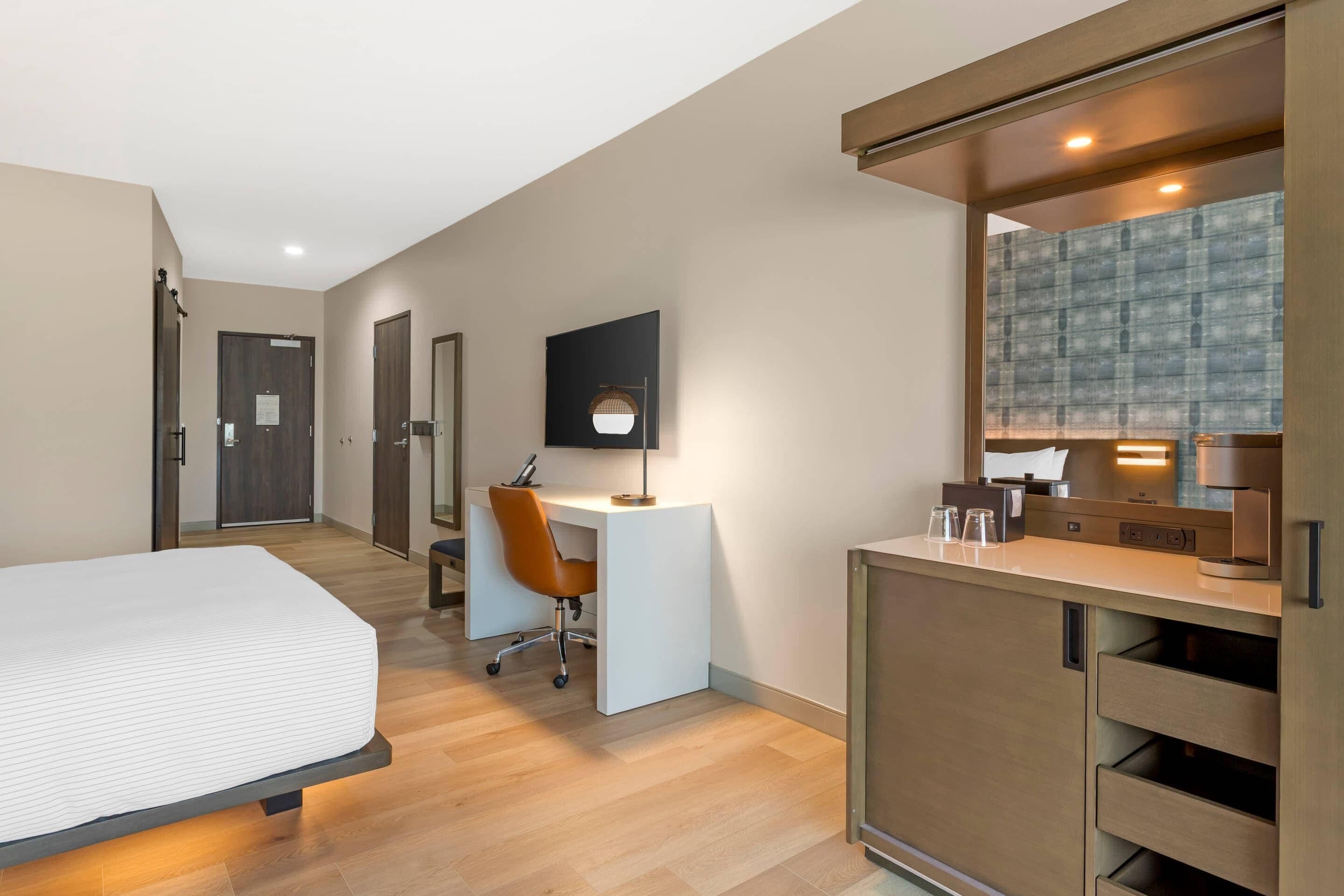 Una habitación de hotel moderna con una cama, un escritorio con una silla, un televisor y un espejo.
