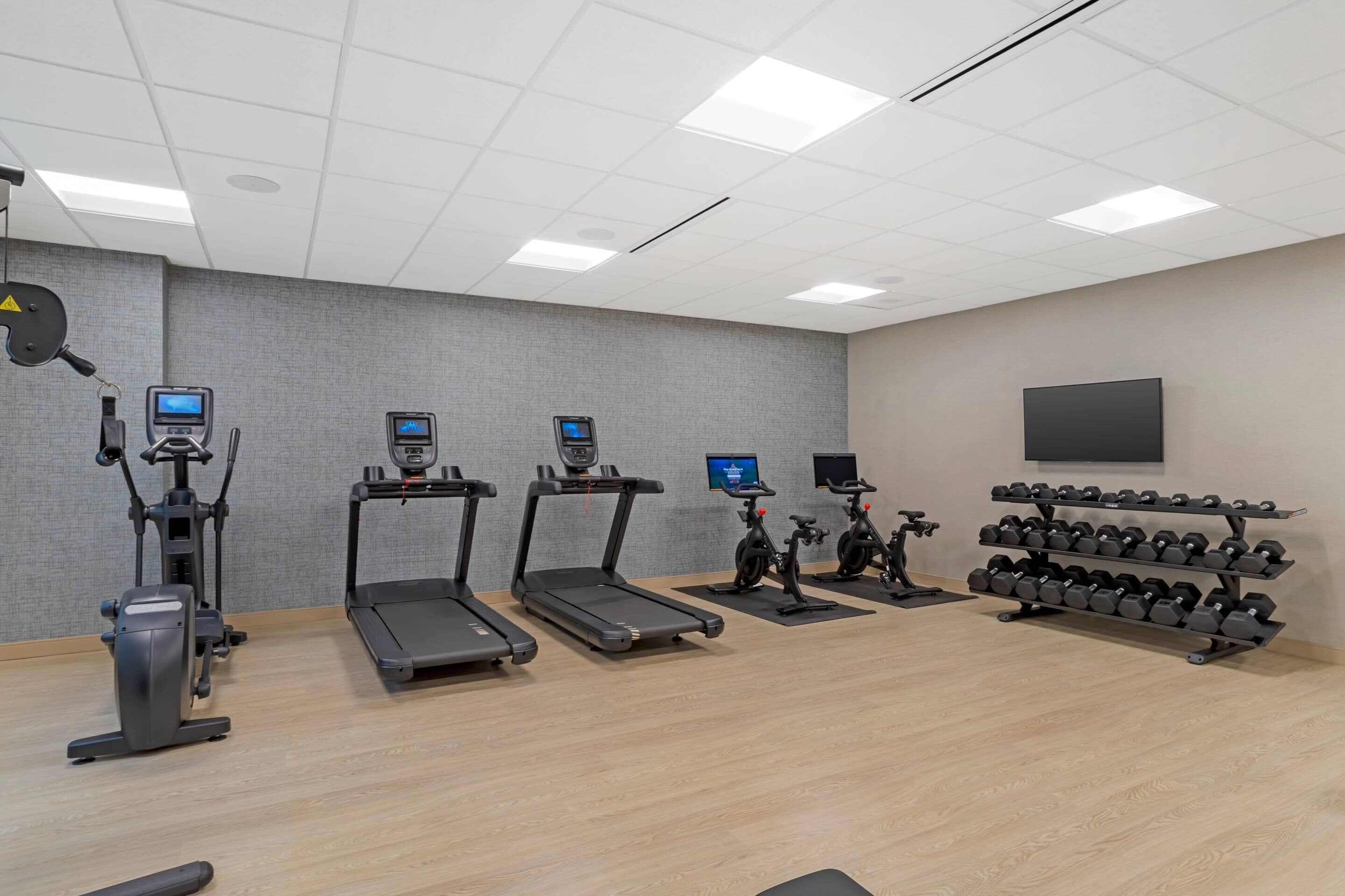 Un gimnasio con cintas de correr, elípticas, bicicletas estáticas y un rack de mancuernas.
