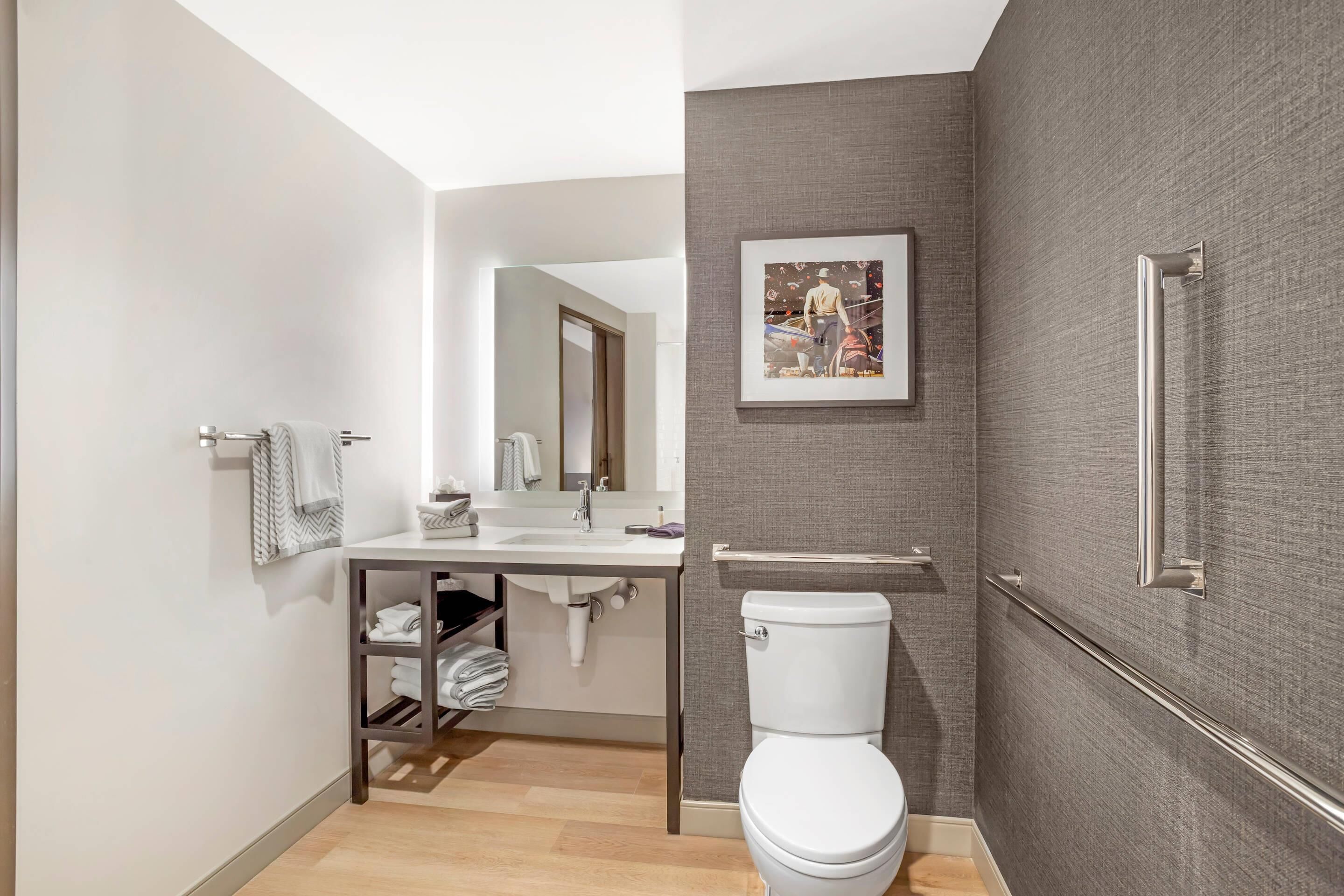 A modern bathroom with a toilet, sink, and mirror. There is a towel rack and a shelf with folded towels.