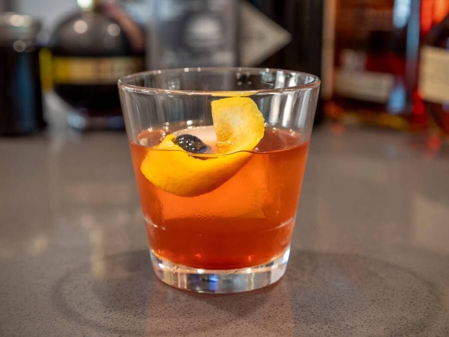 Un vaso transparente con una bebida roja, una rodaja de naranja y un trozo de fruta negra.