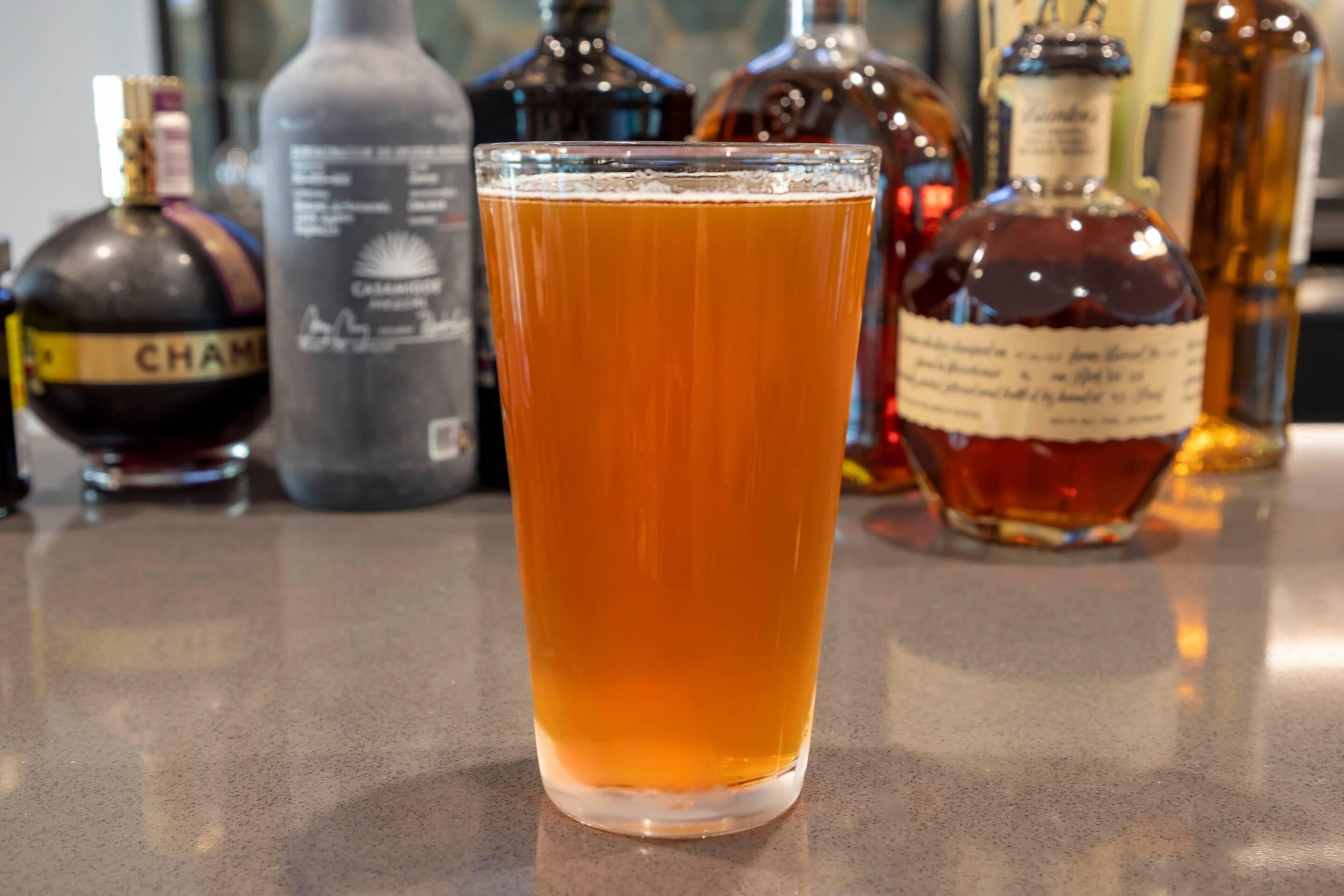 Un vaso de líquido color ámbar, probablemente cerveza, en un vaso transparente. Al fondo, varias botellas de licores diversos, incluyendo una botella con la etiqueta “CHAM”.