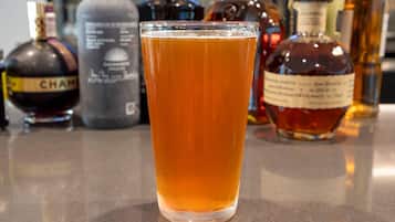 Un vaso de líquido color ámbar, probablemente cerveza, en un vaso transparente. Al fondo, varias botellas de licores diversos, incluyendo una botella con la etiqueta “CHAM”.