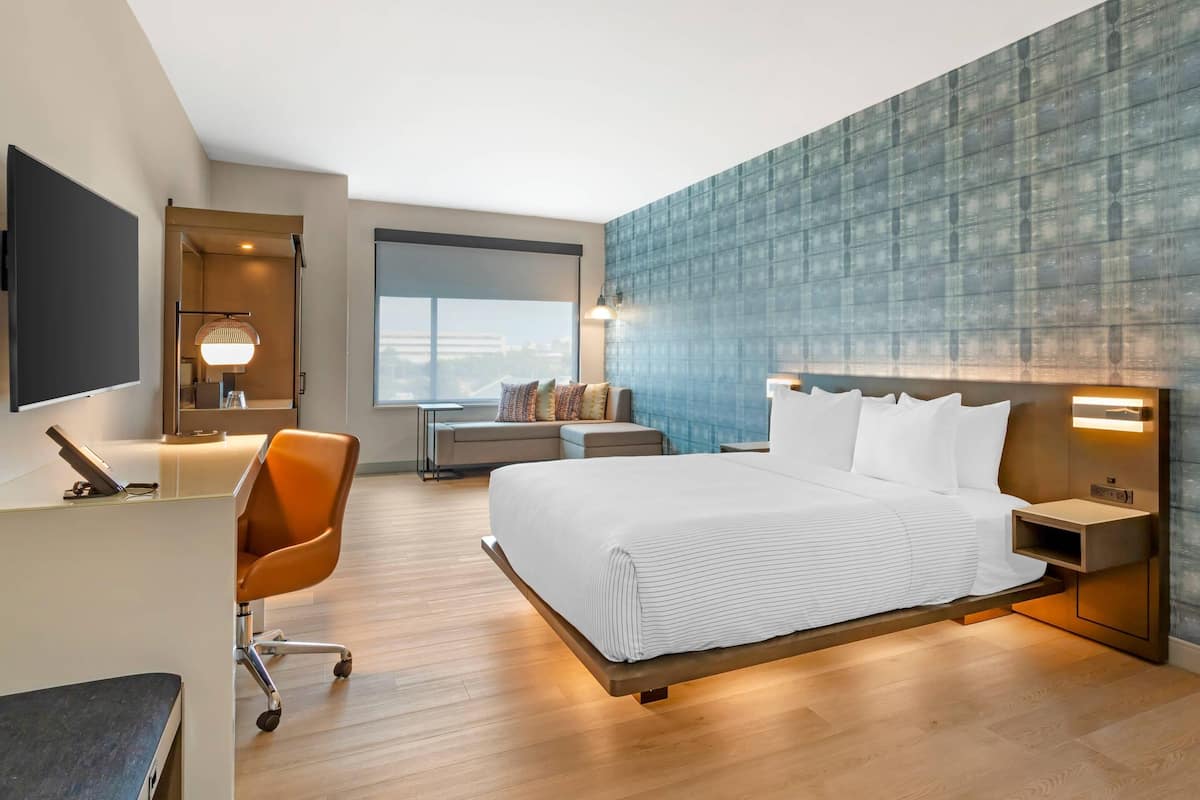 Une chambre d’hôtel moderne avec un grand lit, un bureau, un fauteuil orange et une vue sur la ville.