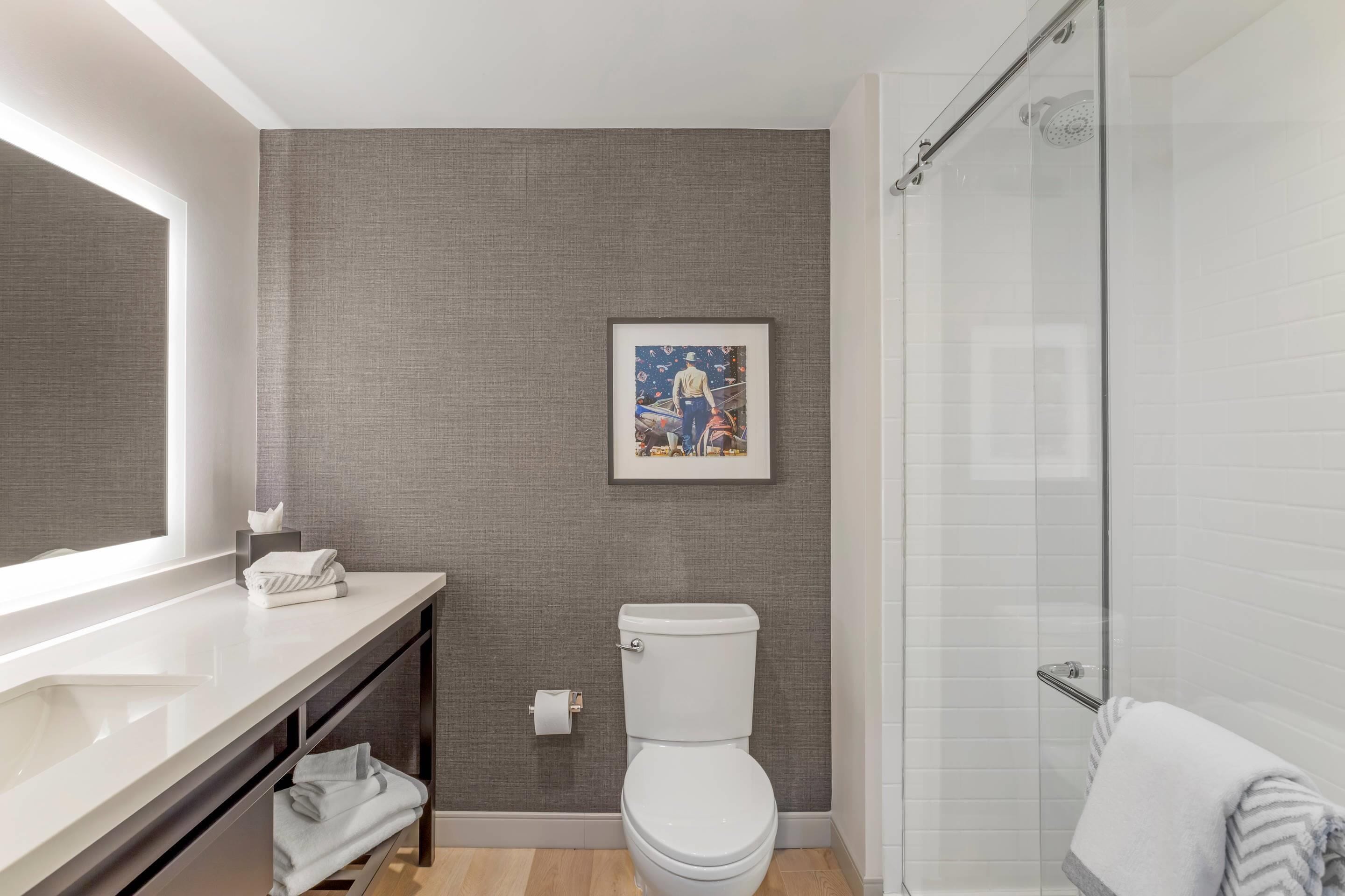 Une salle de bain moderne avec des toilettes blanches, une douche en verre et une œuvre d’art encadrée au mur.