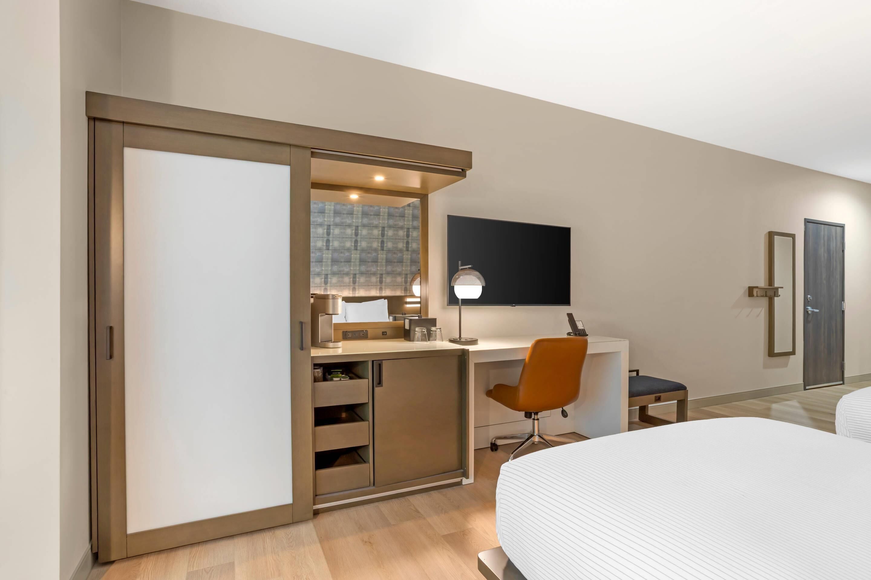 A modern hotel room with a sliding closet, a desk with a chair, a bed, and a TV mounted on the wall.