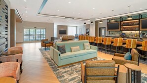 Eine großzügige Lobby mit einem hellblauen Sofa, gemusterten Sesseln, einer Bar mit Hockern und einem großen Bildschirm.