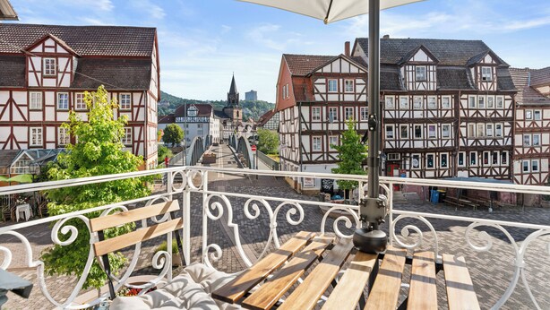 A balcony with a view of traditional half-timbered houses, a cobblestone street, and a church spire in the distance.