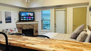 A living room with a long wooden table, a fireplace, a television, and a door leading to a balcony. - Mountain View Studio with Deck, BBQ & Pool (Fernie)