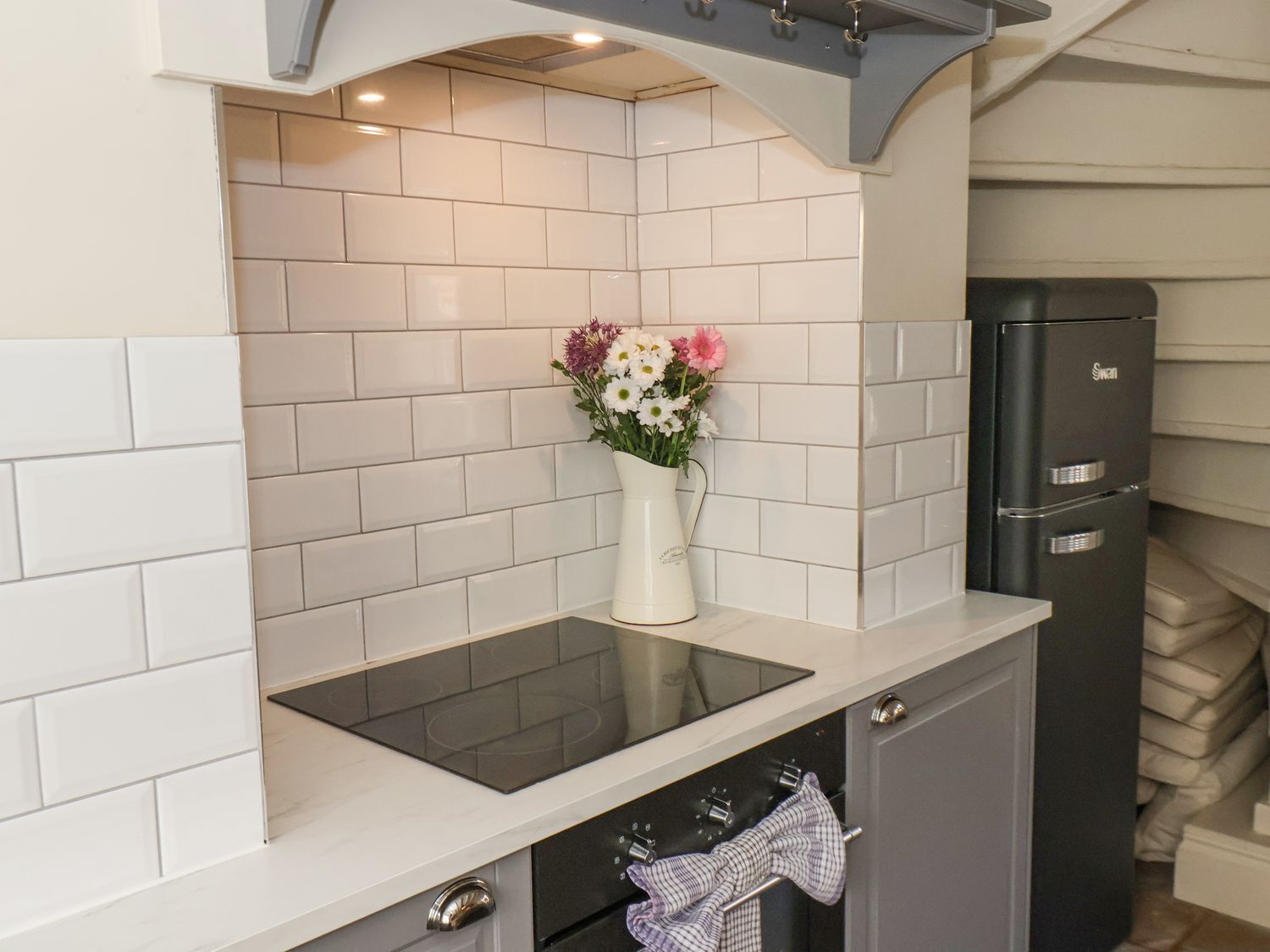 Une cuisine avec des carreaux de céramique blancs disposés en motif métro, une plaque de cuisson à induction noire, un vase blanc orné de fleurs et un réfrigérateur noir.