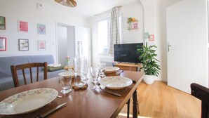 A dining area with a wooden table set for four, a television in the background, and a plant on the right. - LES LOGIS DU GENERAL - Ground floor apartment (Beaupréau-en-Mauges)
