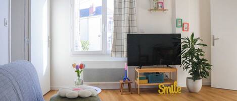 Una sala de estar con un ventanal grande, un televisor de pantalla plana sobre un soporte de madera, una planta en maceta y un letrero que dice 'Smile' en el suelo.