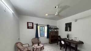 Ein Zimmer mit Marmorboden, Deckenventilator, Esstisch mit Stühlen, Bücherregal und einem Fenster mit Vorhängen.