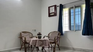 Ein Zimmer mit Esstisch, zwei Korbstühlen, einer Wanduhr und einem Fenster mit Vorhängen.