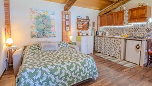 A cozy bedroom with a bed, a nightstand, and a bedside table. Adjacent to the bedroom is a small kitchen with a refrigerator, a sink, and wooden cabinets.