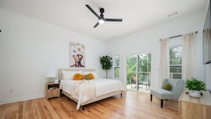 Un dormitorio con cama, mesita de noche, lámpara, ventilador de techo, ventanas amplias y vistas al verdor exterior.