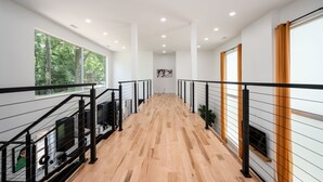 Un interior moderno con piso de madera, barandales negros y amplios ventanales con vistas a árboles.