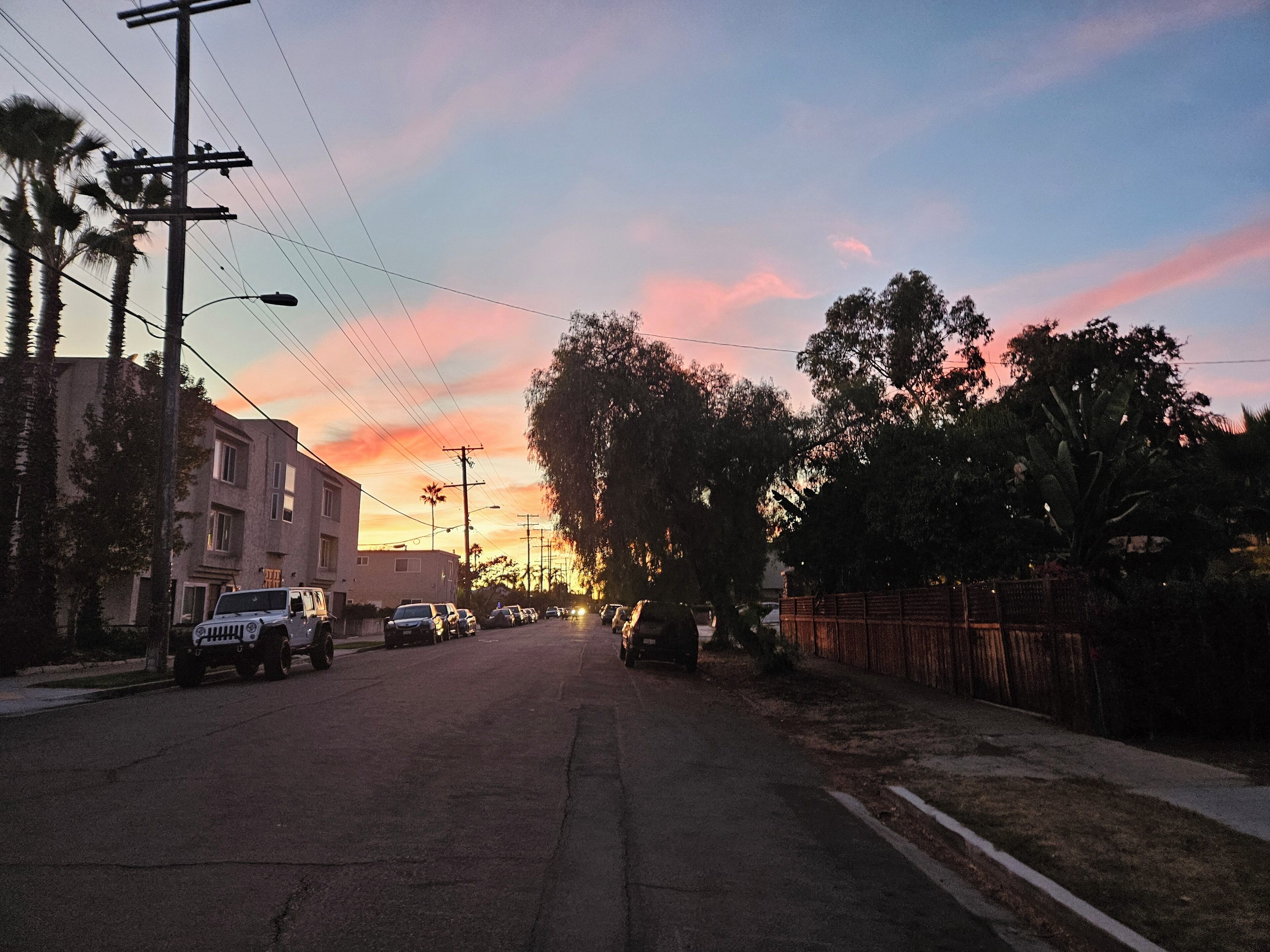 Une vue de rue au coucher du soleil, avec des bâtiments, des voitures garées et des arbres.