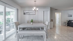 A spacious dining area with a large wooden table and benches, a chandelier, and a view through large sliding glass doors to an outdoor patio. - Charming 2-bedroom house in welcoming Sarasota with AC (Sarasota)