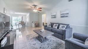 A modern living room with a gray sofa, a coffee table, and a TV. There is a dining area with chairs and a table in the background. - Charming 2-bedroom house in welcoming Sarasota with AC (Sarasota)