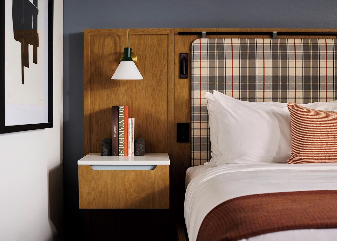 A hotel room with a wooden headboard, a bedside table with a lamp, books, and a bed with white bedding and a brown throw.