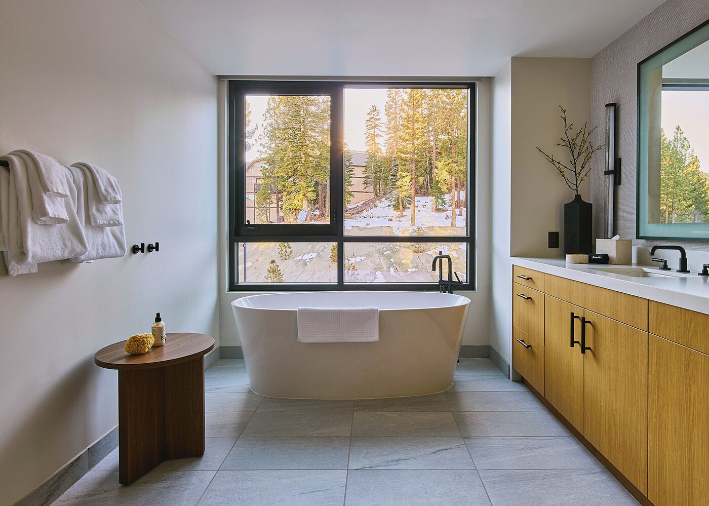 A modern bathroom with a freestanding bathtub, a vanity with a mirror, and a large window offering a view of trees and a building.