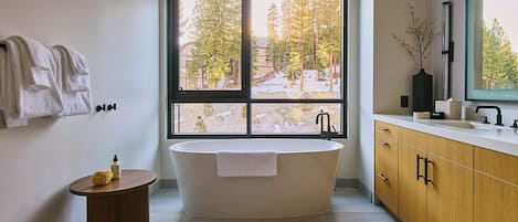 Un baño moderno con una bañera independiente, un tocador con espejo y un amplio ventanal que ofrece vistas a árboles y un edificio.