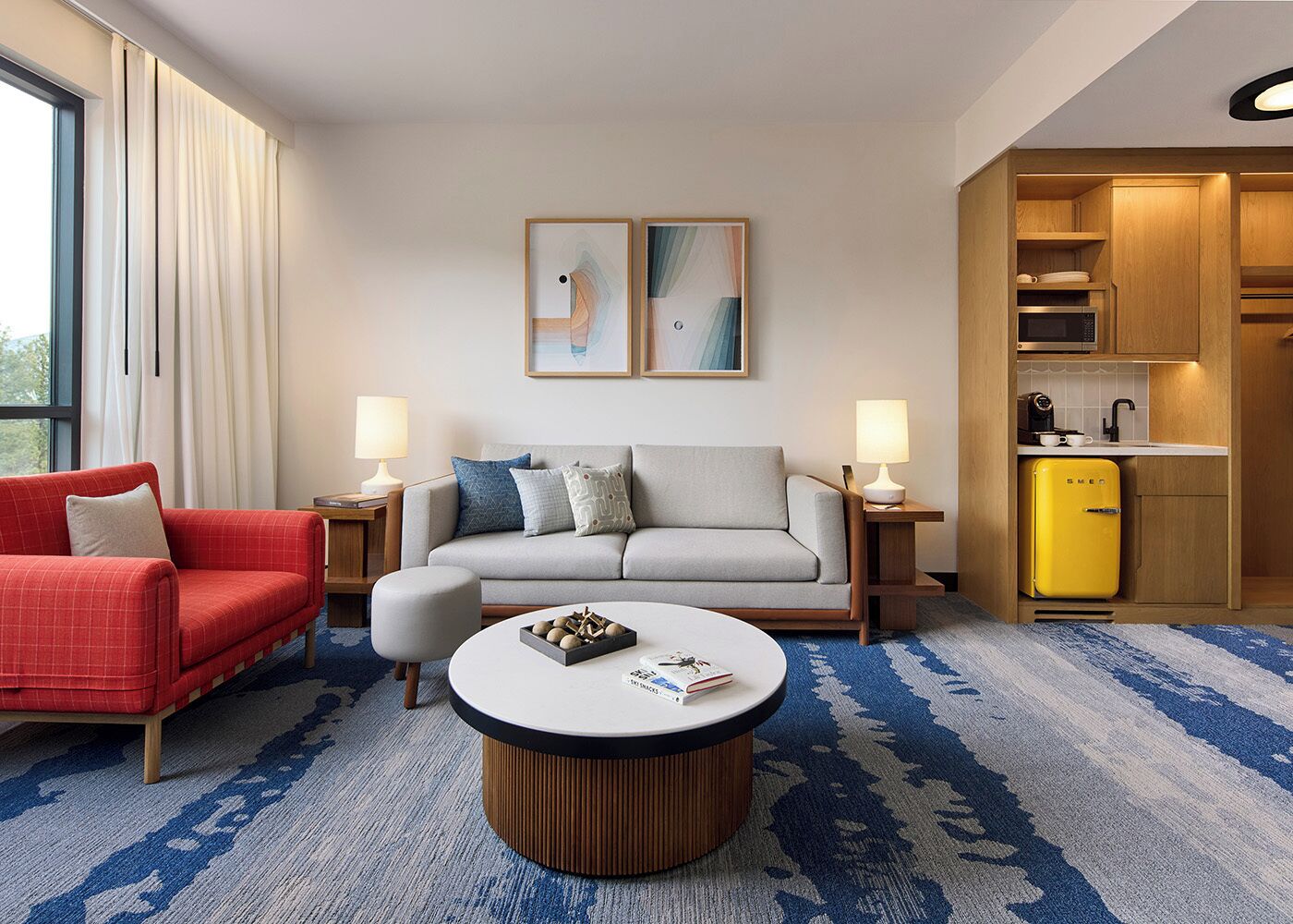 A hotel room with a red armchair, grey sofa, and a small round table with a book. There are two wall-mounted lamps, a microwave, and a yellow refrigerator in the kitchen area.
