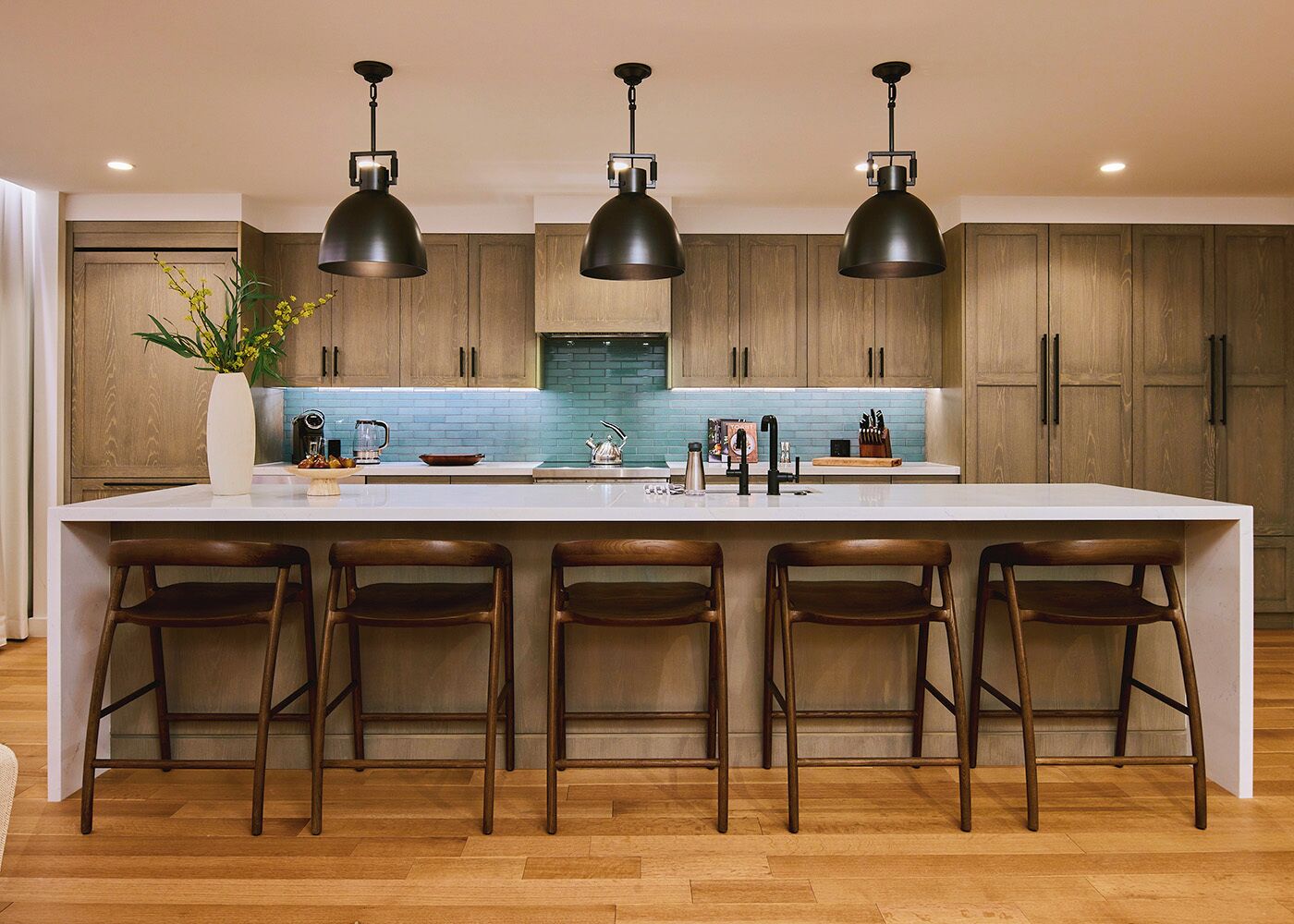 A modern kitchen with a large island, bar stools, pendant lights, and wooden cabinets.