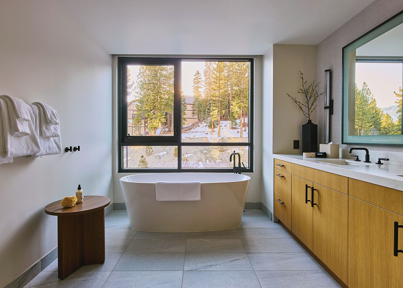 A modern bathroom with a freestanding bathtub, a large window with a view of trees, a wooden vanity, and a small round table with a bottle.