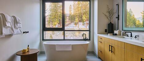 A modern bathroom with a freestanding bathtub, a large window with a view of trees, a wooden vanity, and a small round table with a bottle.