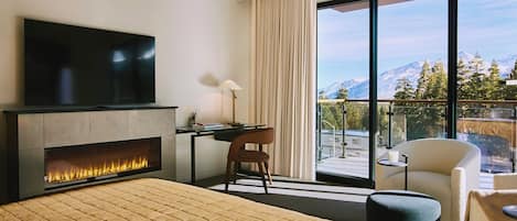 A hotel room with a bed, a fireplace, a desk, a chair, a TV, and a balcony with a mountain view.