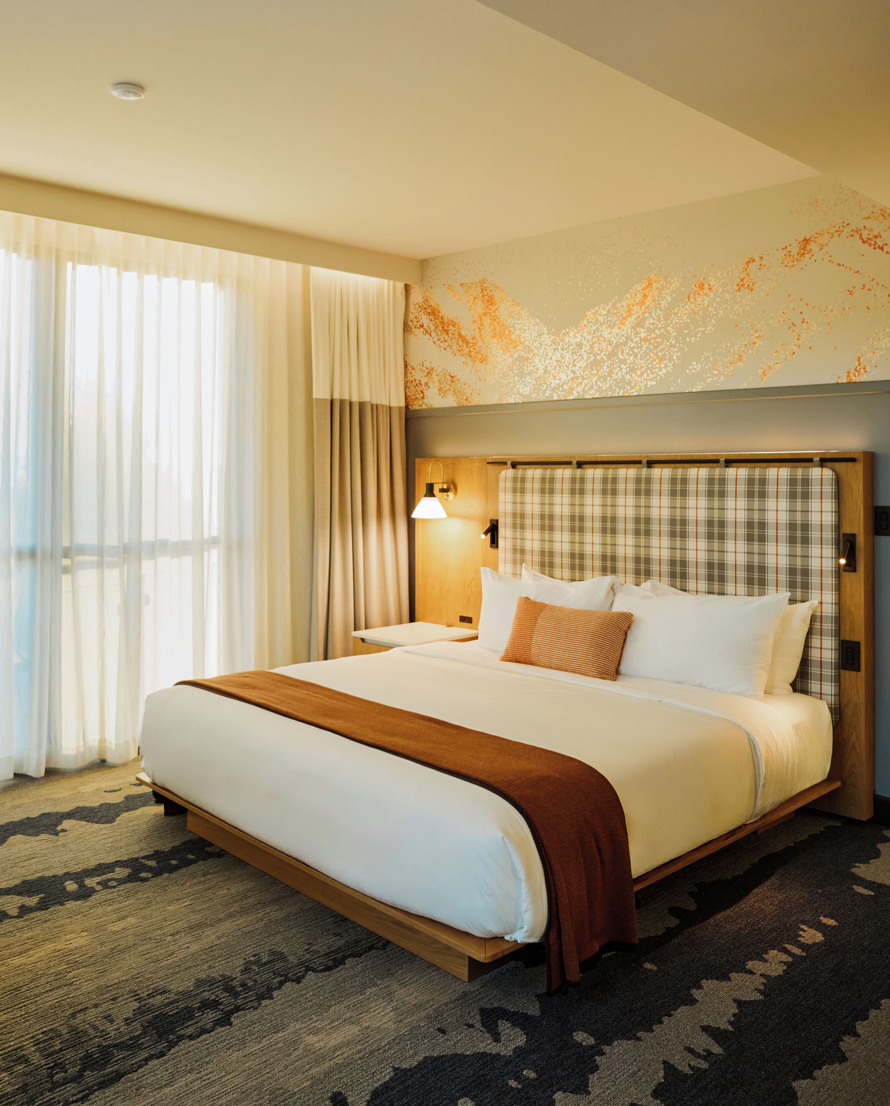 Una habitación de hotel con una cama grande, una mesita de noche, una lámpara y una ventana con cortinas.