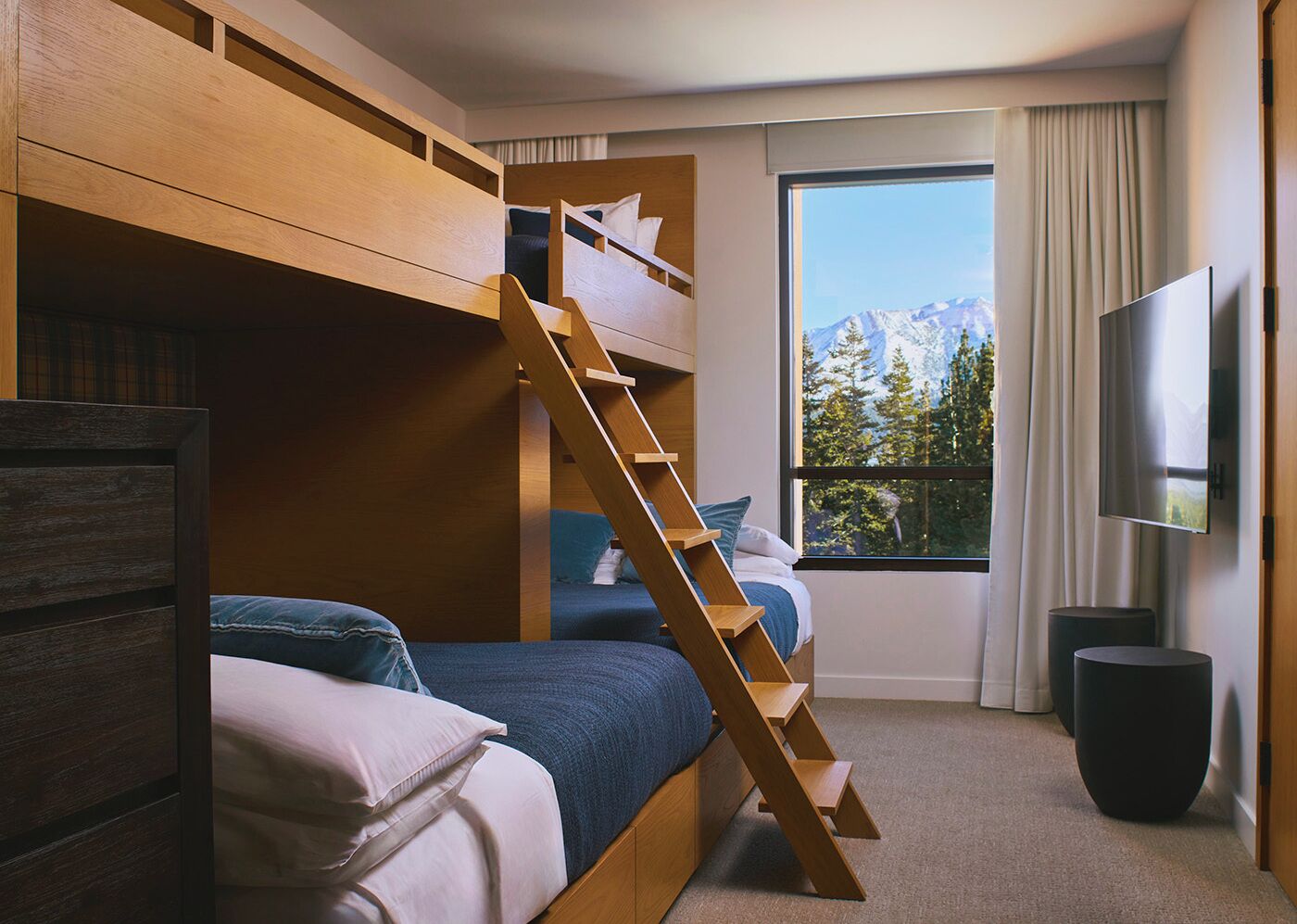 A room with bunk beds, a wooden ladder, a TV, and a window with a mountain view.