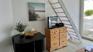 A room with a wooden cabinet, a black table with a plant, a painting of a beach, and a staircase leading to a balcony.