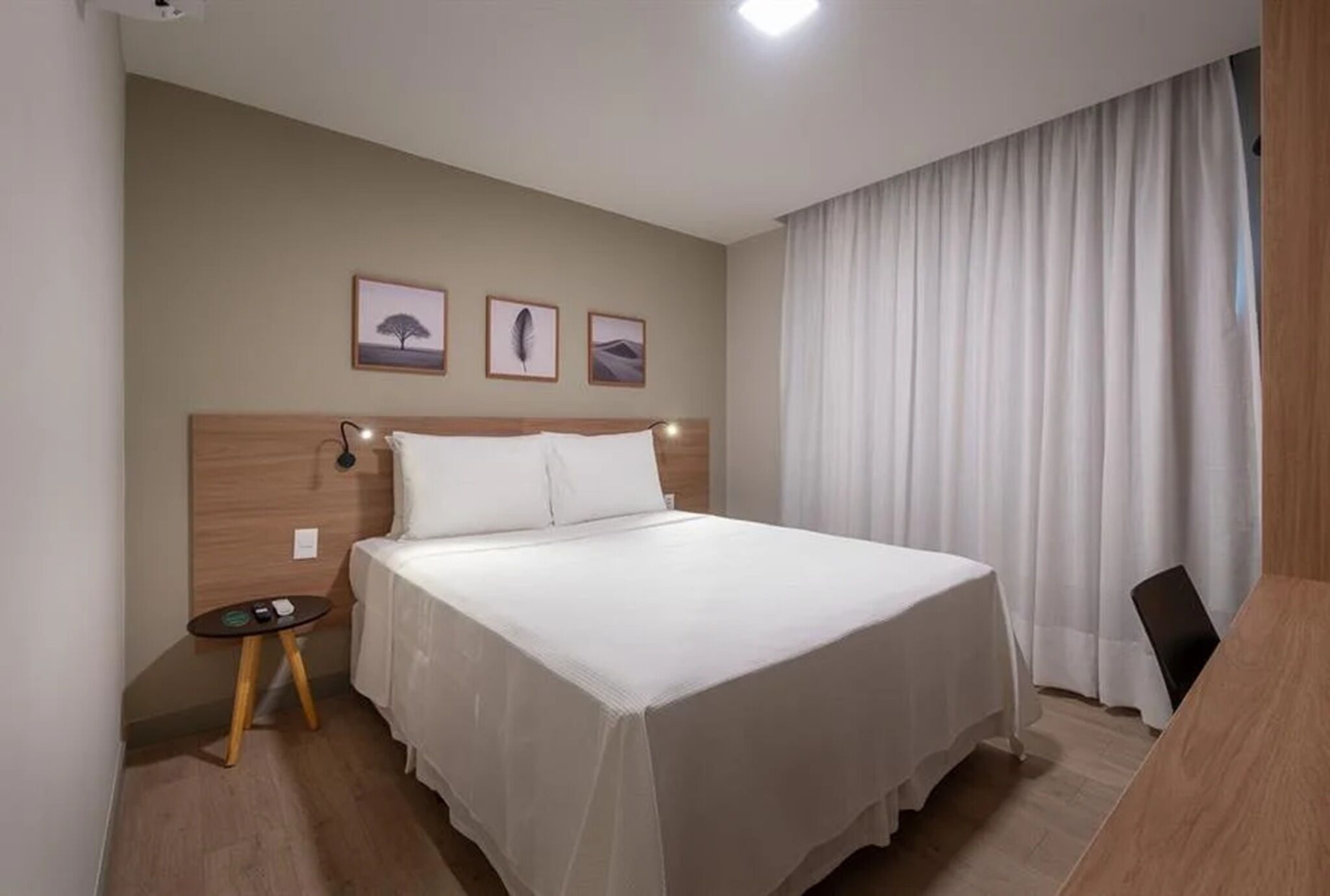A hotel room with a bed, a small table, a chair, and three framed pictures on the wall.