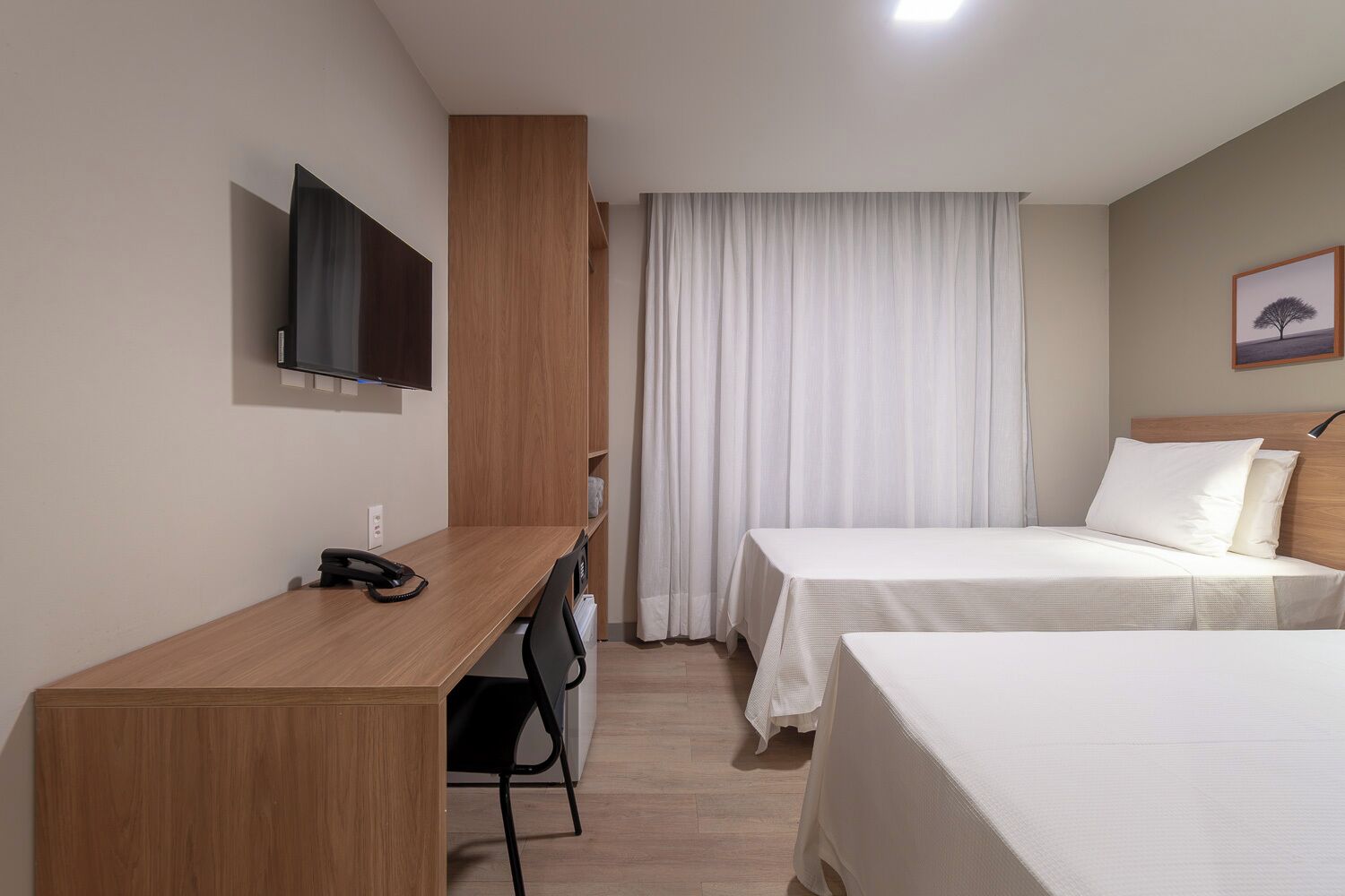 A hotel room with a wooden desk, a bed, a TV mounted on the wall, and a window with curtains.