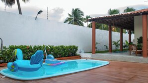 Una piscina con juguetes inflables, un área de estar techada y una pared blanca con plantas.