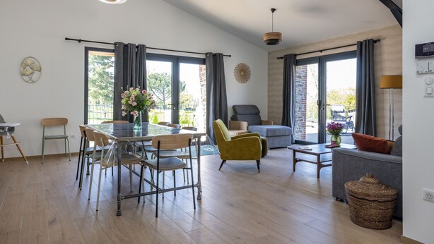 A spacious living area with a dining table, chairs, a sofa, and a coffee table. There are large windows with curtains, a lamp, and a wall-mounted thermostat. - Les Genêts - PMR gîte - pets welcome (Saint-Christophe-du-Bois)