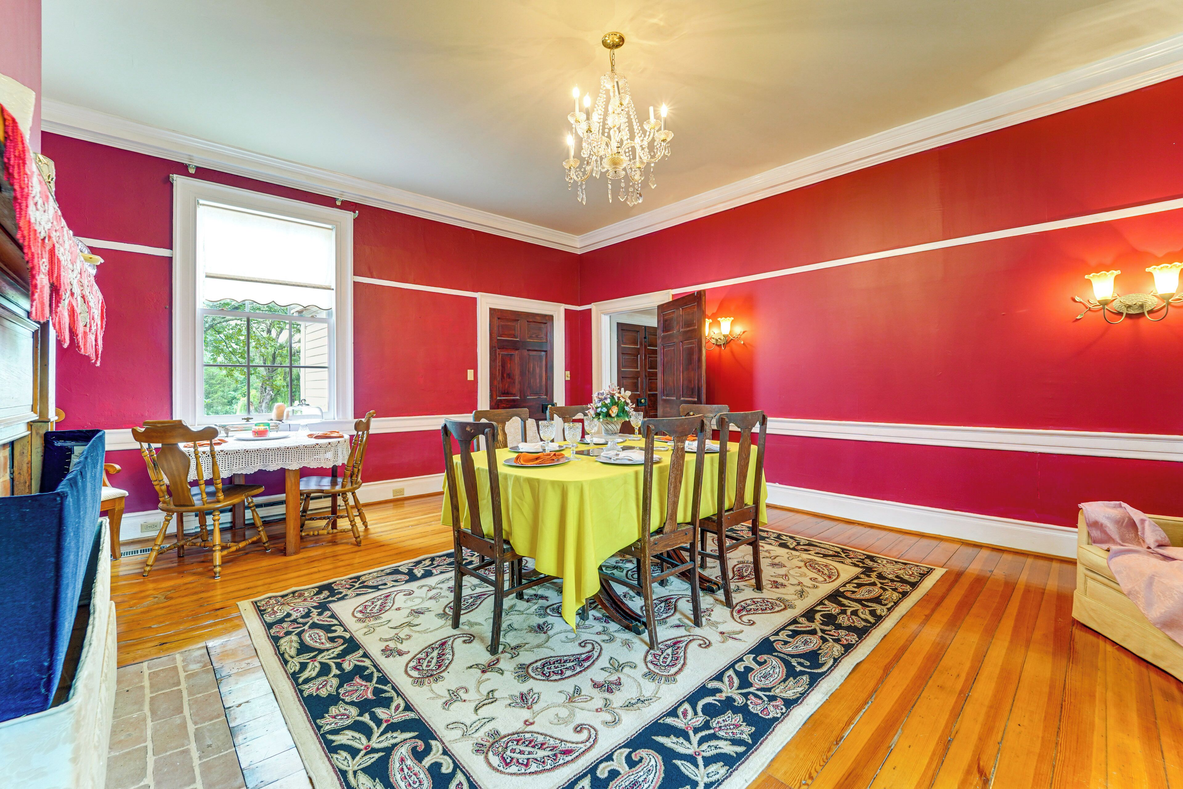 Uma sala de jantar com parede vermelha, um candelabro, uma mesa de madeira com cadeiras e um tapete estampado.