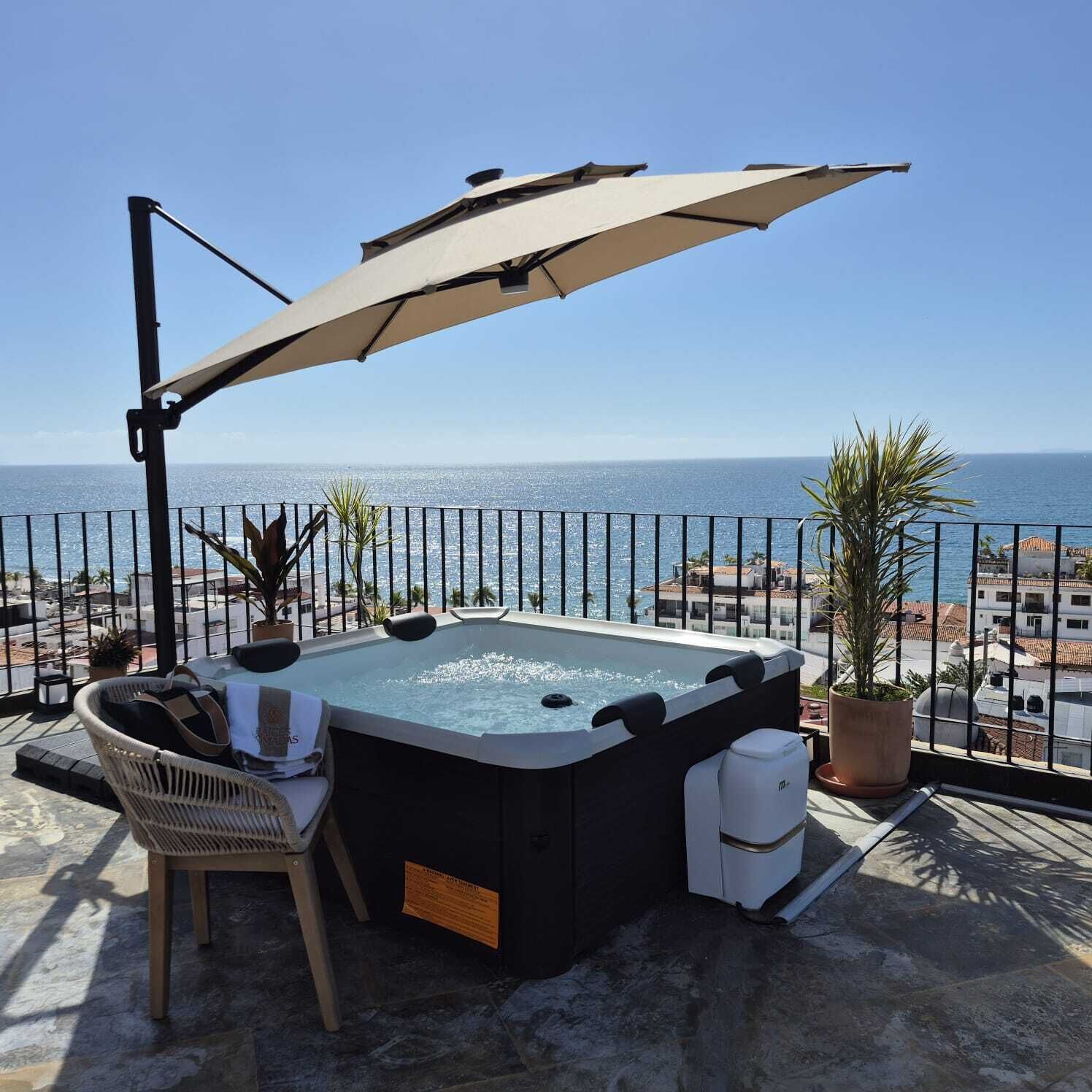 A hot tub with a view of the sea, a chair, a potted plant, and an umbrella.