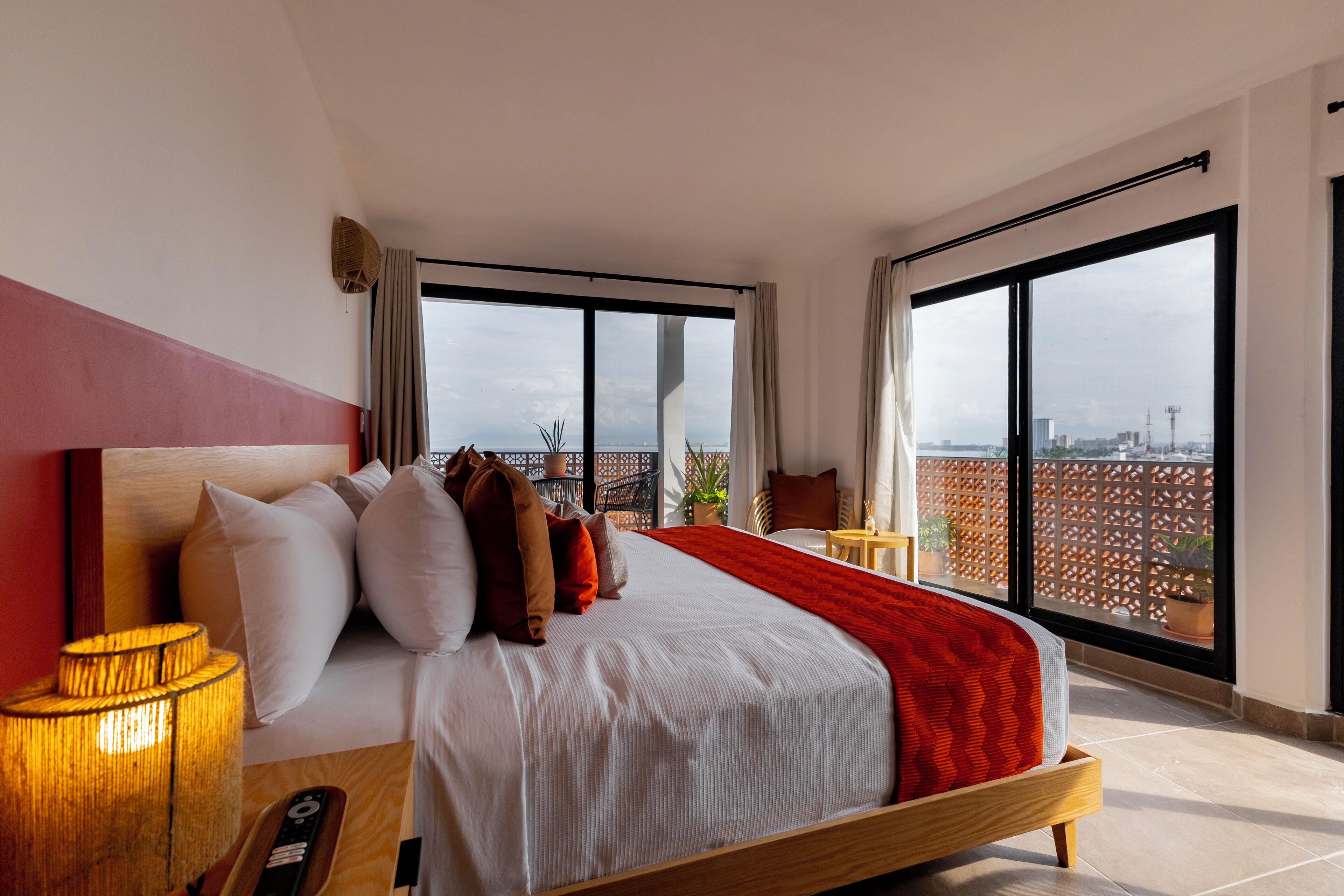 A modern hotel room with a large bed, a bedside table, a lamp, and a view of the city through large windows.