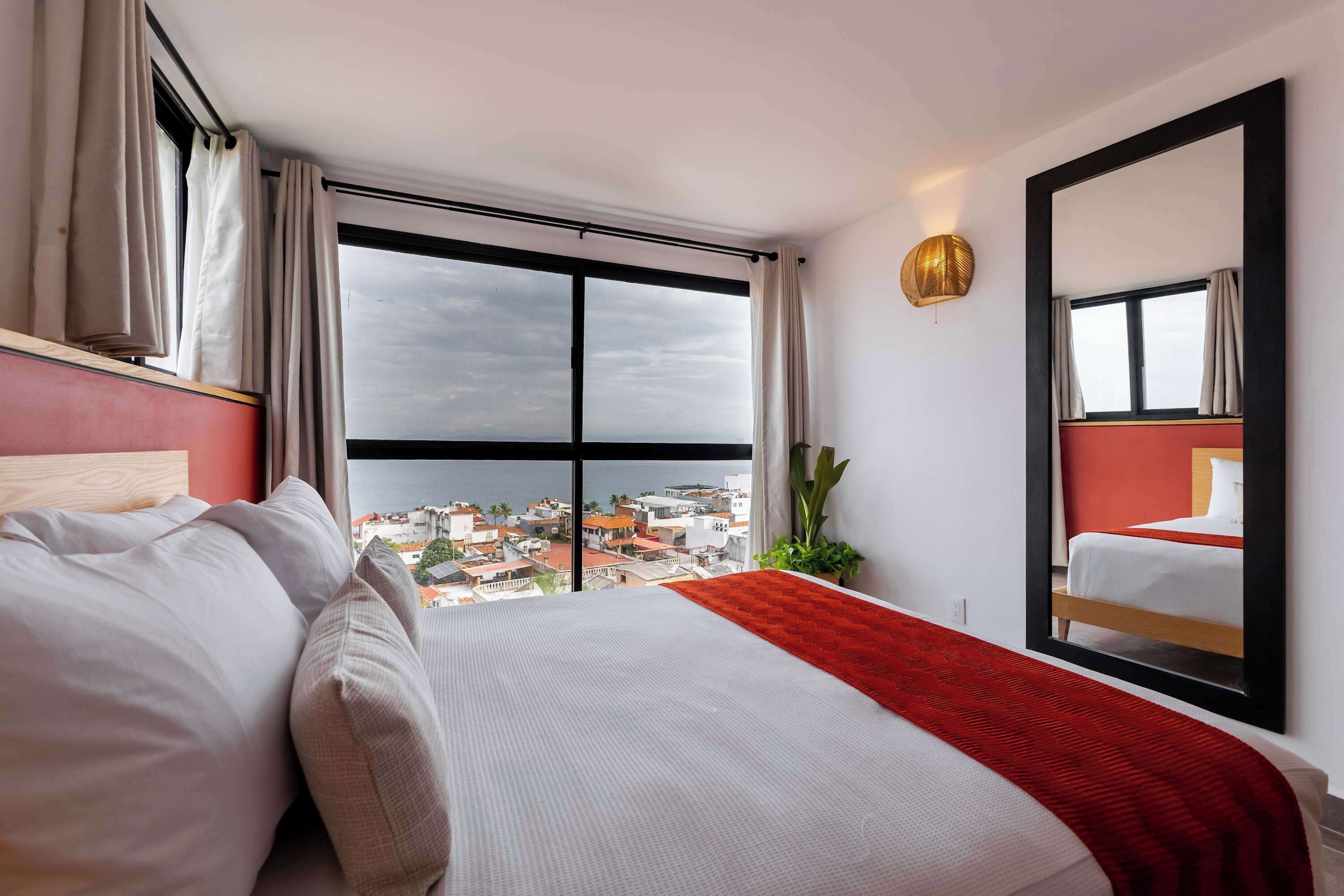A hotel room with a large window, a bed with white bedding, a red throw, and a mirror on the wall.